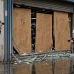 Devanture d’un magasin de sport avec vitrine endommagée et protégée par des panneaux, tandis qu’une femme et un homme réalisent un constat photographique.